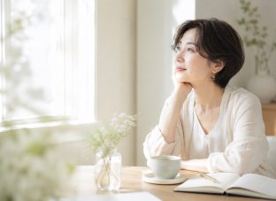 窓辺のやわらかな光の中で、ノートとカップを前に静かに考えごとをする40代50代女性