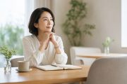 明るい室内のテーブルで、開いたノートを前に穏やかに考える女性