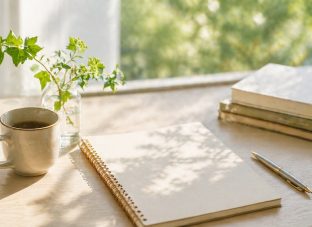 明るい日差しが入る窓辺の木のテーブルに、ノート、ペン、湯気の立つマグカップ、小さなグリーンの花瓶、重ねた本が静かに置かれた、やわらかく落ち着いたイメージ画像。
