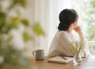 やわらかな自然光が入る窓辺で、ノートとカップを前に静かに外を眺める女性。仕事や人間関係の責任を一人で抱え込まず、自分に合う形に整えていく時間をイメージしたアイキャッチ画像。