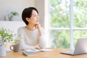 アラフィフ女性が、明るい室内のデスクでノートとパソコンを前に、窓の外を見ながらこれからの働き方を考えている様子