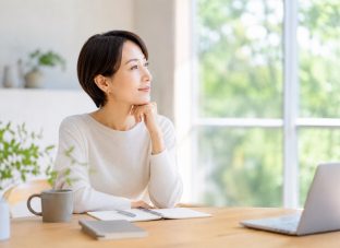 アラフィフ女性が、明るい室内のデスクでノートとパソコンを前に、窓の外を見ながらこれからの働き方を考えている様子