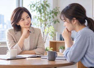 明るいオフィスで、丸テーブルを挟んで向かい合い、穏やかに対話する二人の女性。片方がやさしく話を聴き、もう片方が考え込みながら相談している様子。