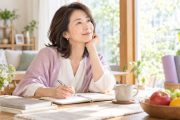 明るい自宅でノートを開き、これからの仕事や家族、体調を含めた暮らし方について考える40代後半から50代の女性。