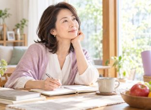 明るい自宅でノートを開き、これからの仕事や家族、体調を含めた暮らし方について考える40代後半から50代の女性。